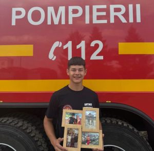 Tânăr zâmbind, ținând un colaj de fotografii, în fața unei autospeciale de pompieri.