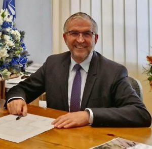 Servicii medicale și sprijin la domiciliu pentru bunicii din Comănești: Înscrierile încep din 21 septembrie 2 Bărbat zâmbitor în costum, la birou, cu flori în fundal.