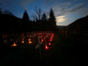 Lumânări aprinse lângă cruci în cimitir, în amurg.