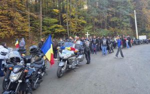 Participanți la omagierea militarilor, cu motociclete și drapeluri românești lângă un drum de pădure.