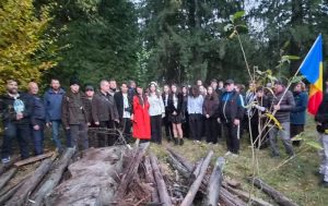 Mulți oameni adunați în pădure lângă cilindrii și trunchiuri de copac la un eveniment patriotic.