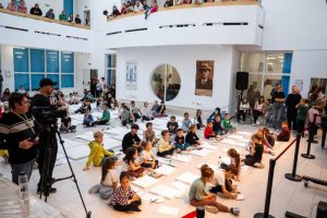 Copii desenând pe podea într-un spațiu public modern, cu spectatori la etaj.