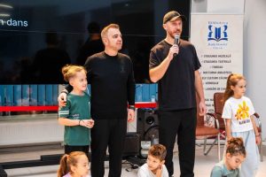 Grup copii pe scenă la eveniment cultural, cu un vorbitor și organizatori în fundal.