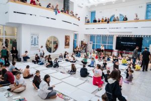 Copii desenând pe pânze albe într-un spațiu amplu cu vizitatori la etaj.
