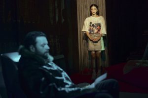 Tânără pe trepte privește într-un spațiu interior întunecat, bărbat așezat în fața unui scaun.