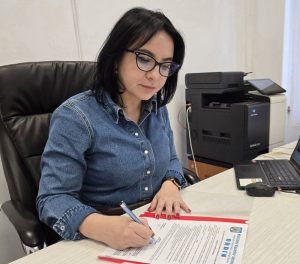 Femeie care semnează un document la birou, înconjurată de echipamente de birou.